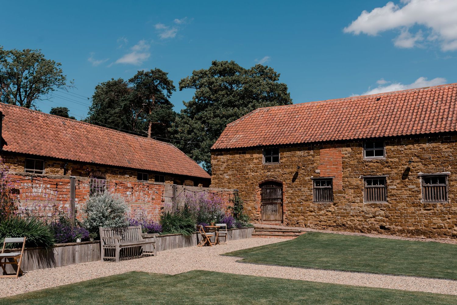 Rustic stone and brick farm buildings with terracotta roofs sit beside a manicured lawn on a sunny day.