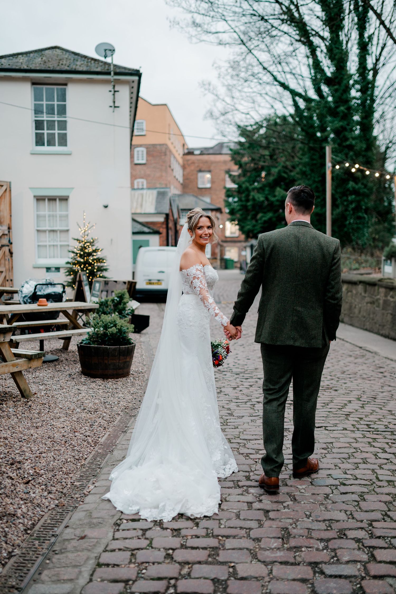 Bride and groom walking hand in hand on cobblestone street, bride in lace wedding gown with train, groom in dark suit.