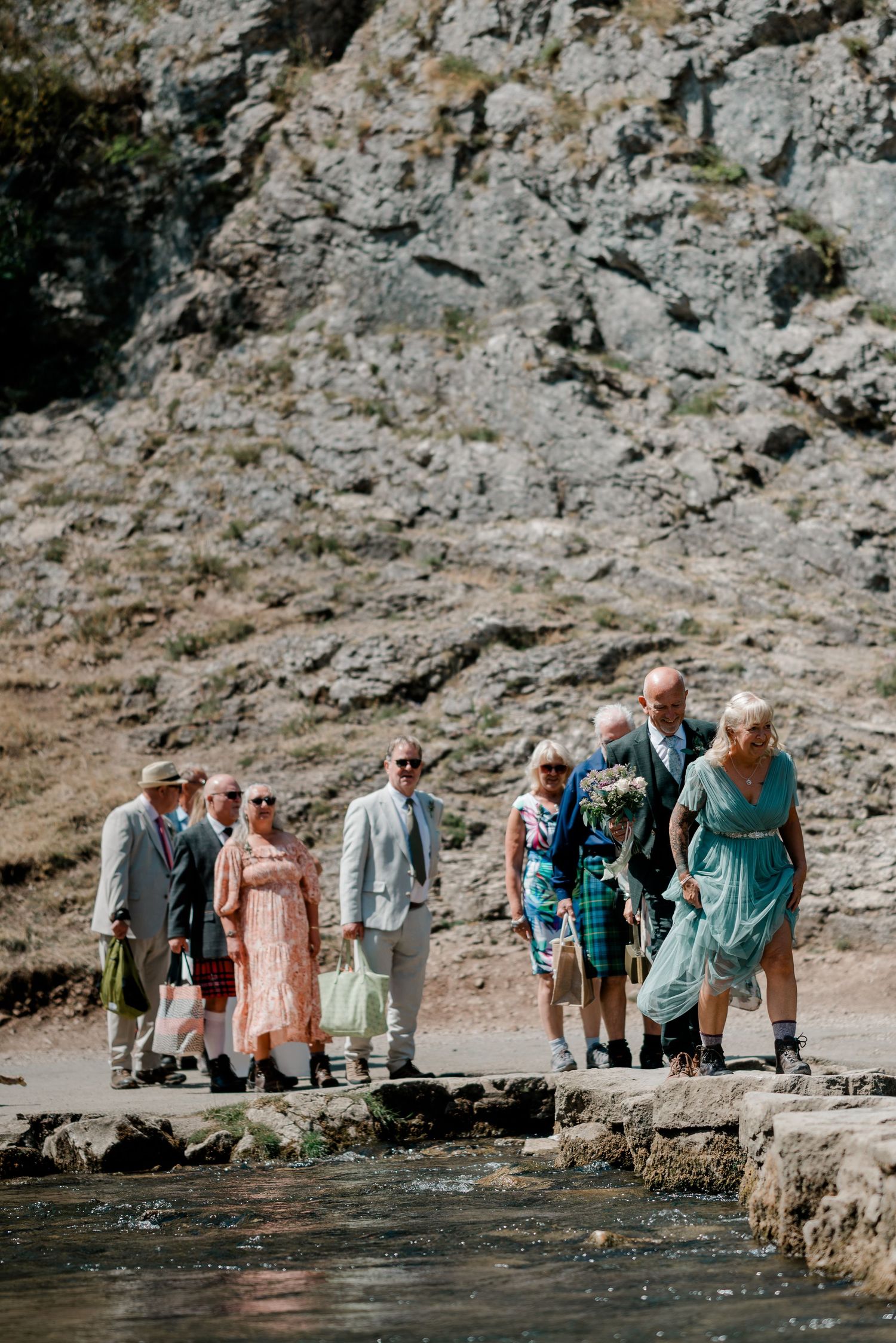 Wedding party crossing stepping stones by a rocky cliff, bride holding bouquet in colorful dress, guests in formal attire outdoors.