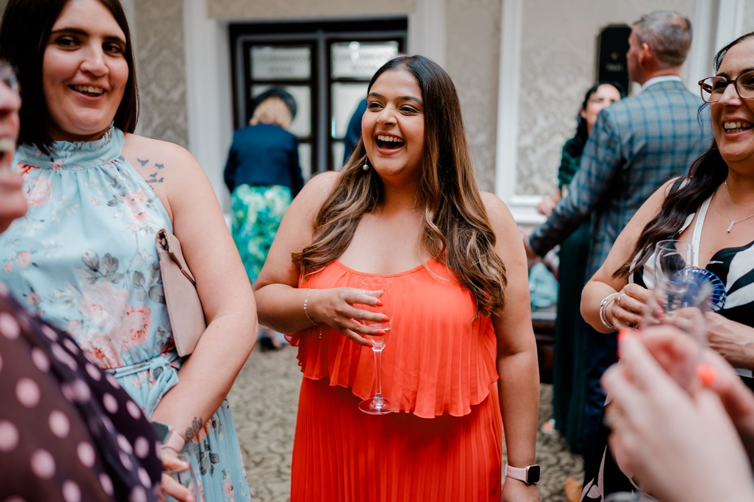 Woman in red dress laughing at outdoor wedding reception holding champagne glass.
