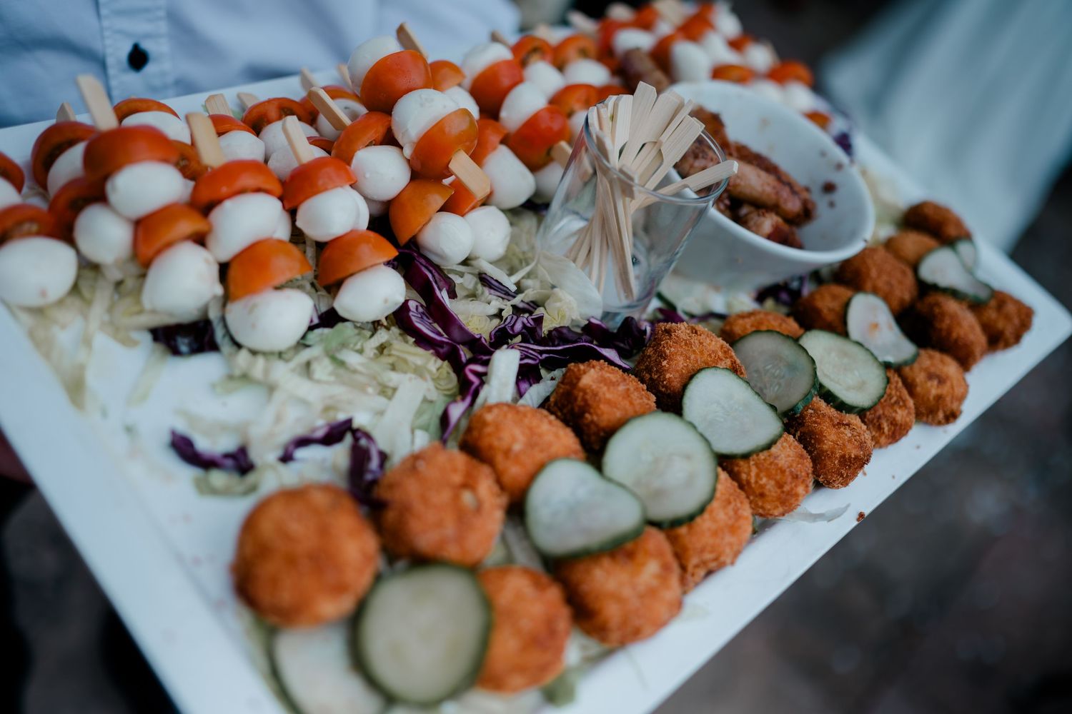 Catered appetizer platter with mozzarella tomato skewers, crispy fried bites, sliced pickles, and shredded red cabbage on a white tray.