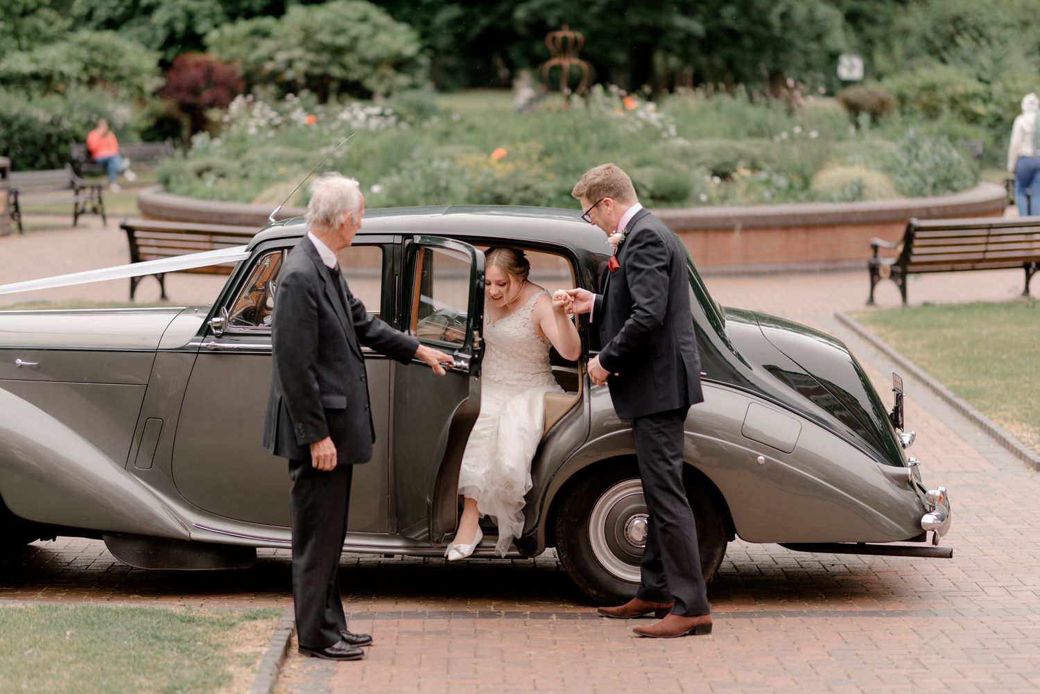 Bride in white dress stepping out of vintage gray car assisted by two men in suits at outdoor wedding venue.