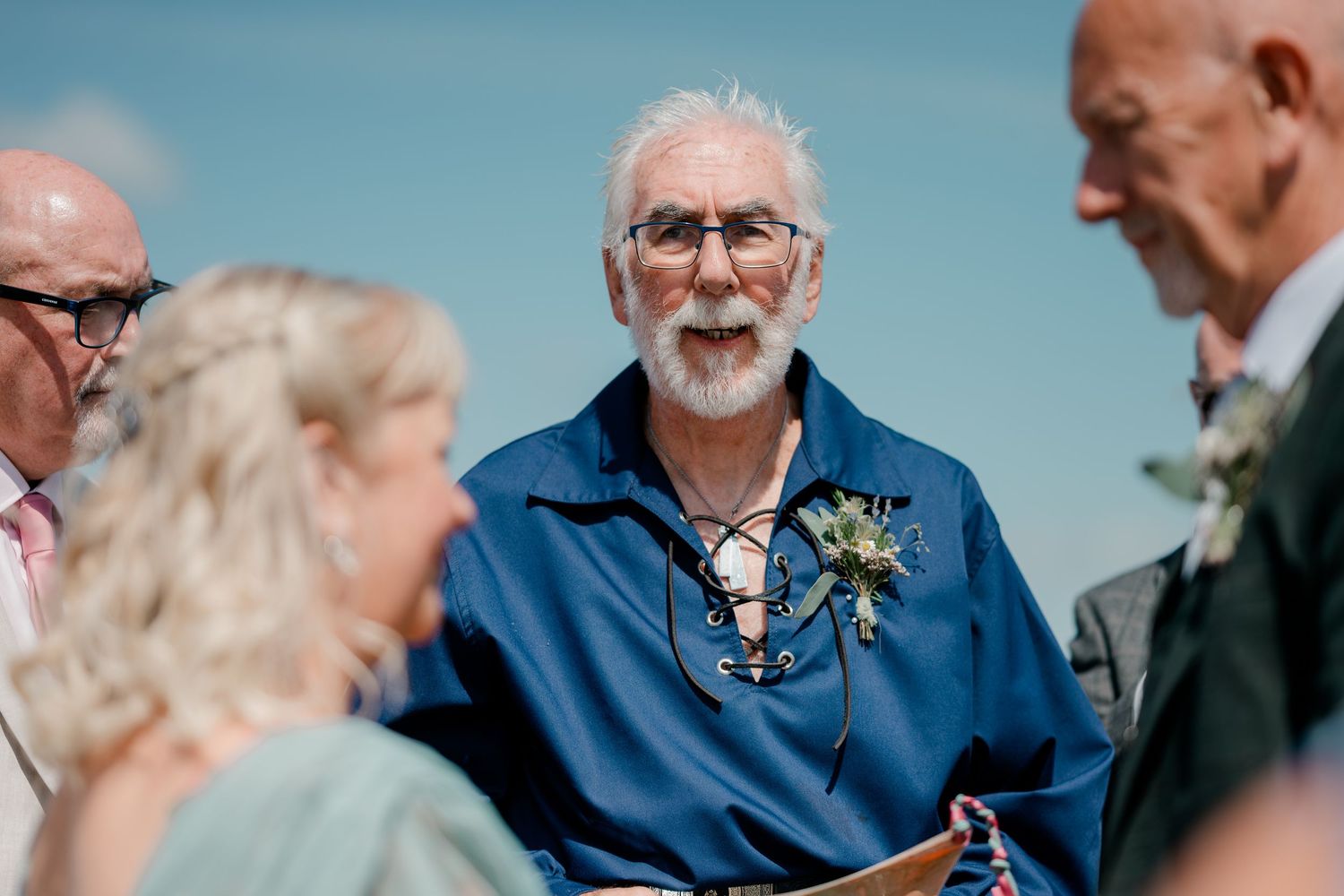 An elderly officiant with white hair speaks during an outdoor wedding ceremony on a sunny day.