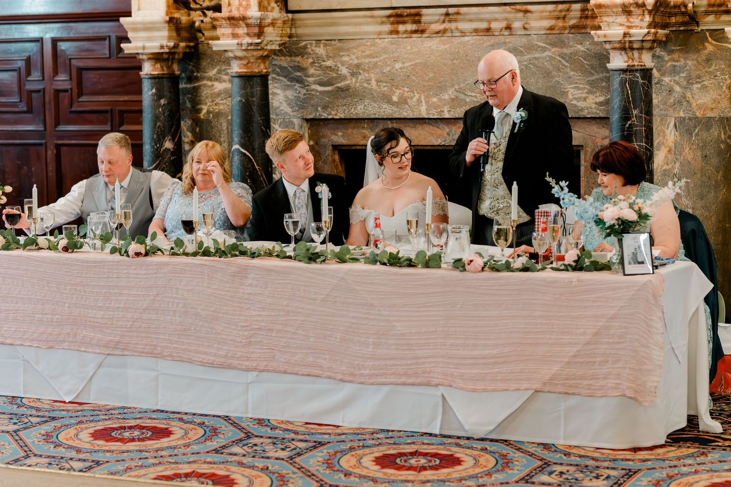 Wedding party seated at head table during reception speech, elegant venue with floral centerpieces and ornate fireplace backdrop.