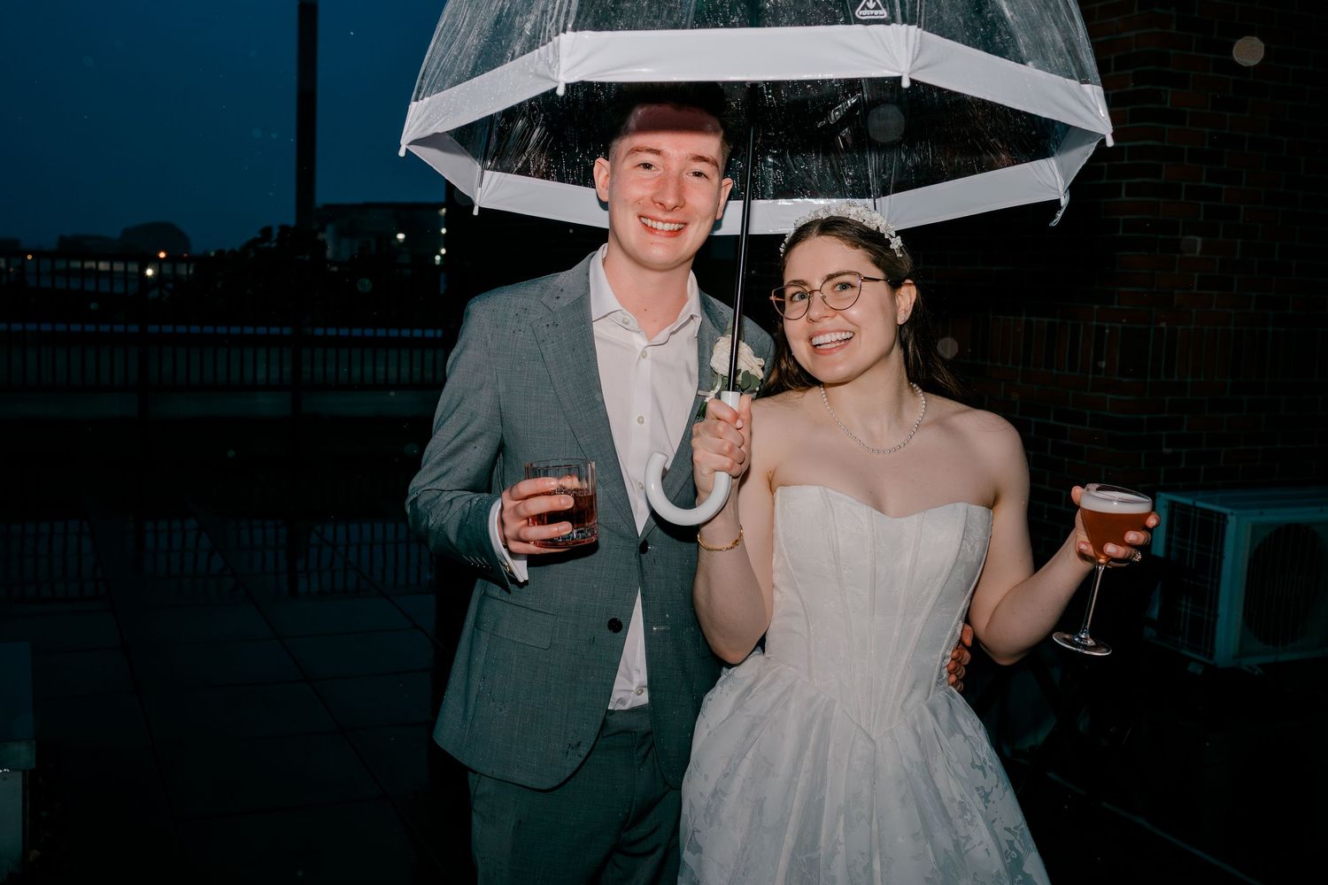 Bride and groom smiling under a clear umbrella outdoors at night, each holding a drink.