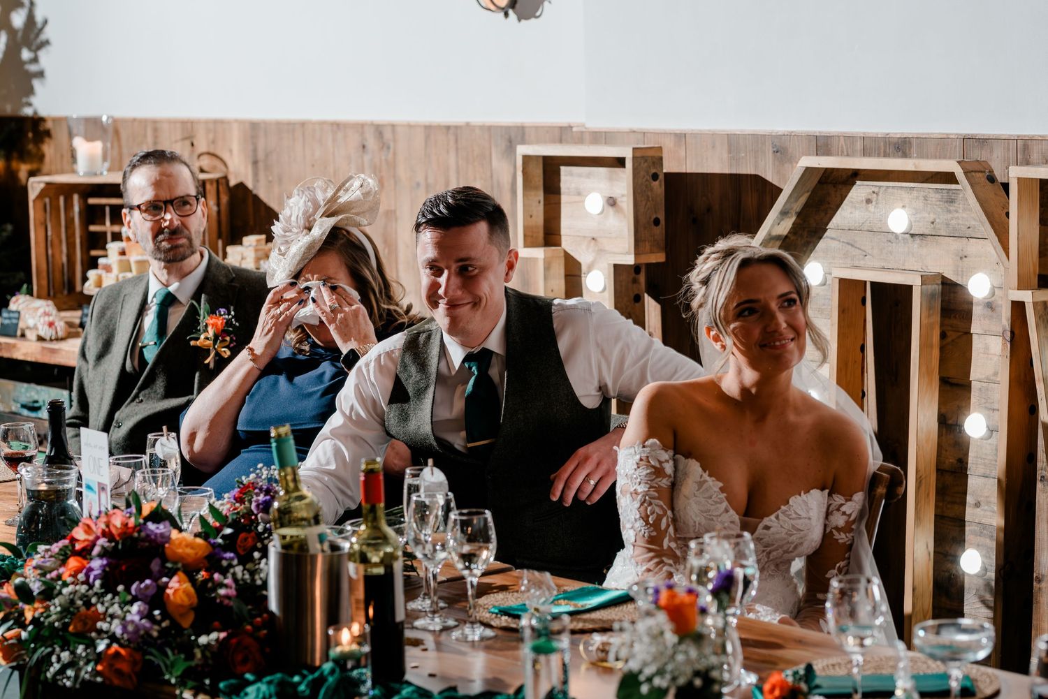 Wedding party laughs and cries during emotional speeches at a rustic barn reception.