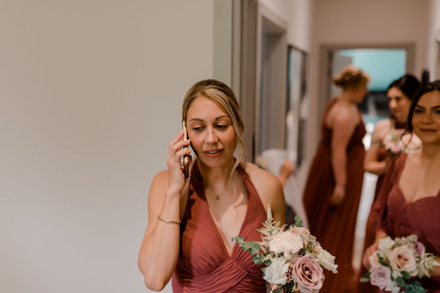 Bridesmaid in dusty rose gown talks on phone while holding bouquet, with other bridesmaids in background.
