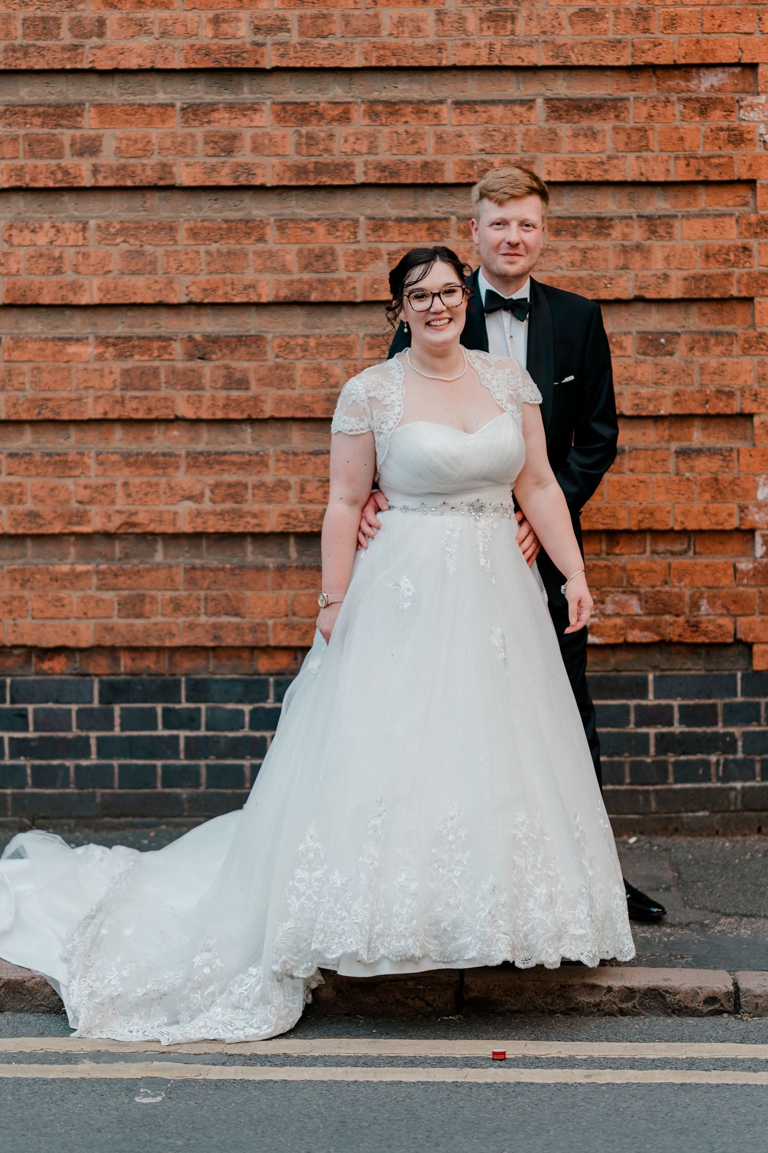 Bride in white lace gown and groom in black tuxedo posing together in front of a red brick wall.
