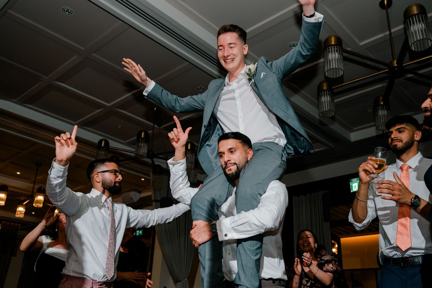 Groom lifted on shoulders by groomsmen celebrating on wedding reception dance floor in elegant venue.