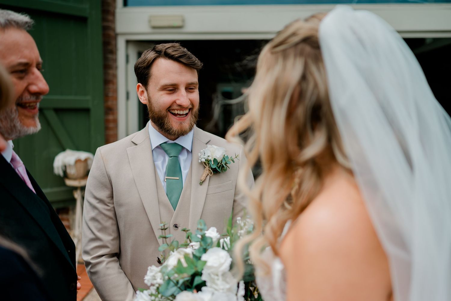 Groom in tan suit smiling joyfully at bride during outdoor wedding ceremony with white floral bouquet.