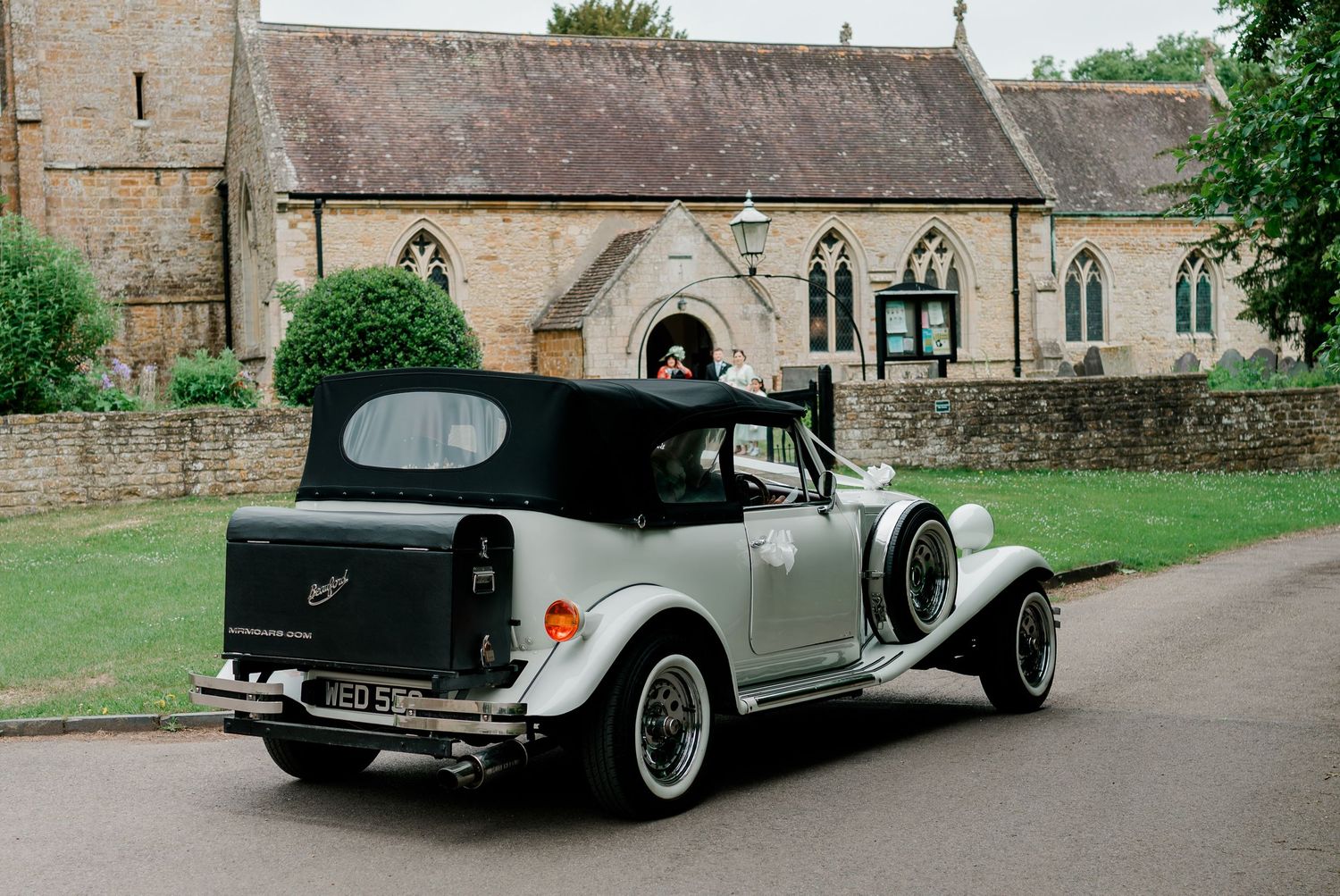 Vintage white and black convertible wedding car parked outside a historic stone church on a sunny day.