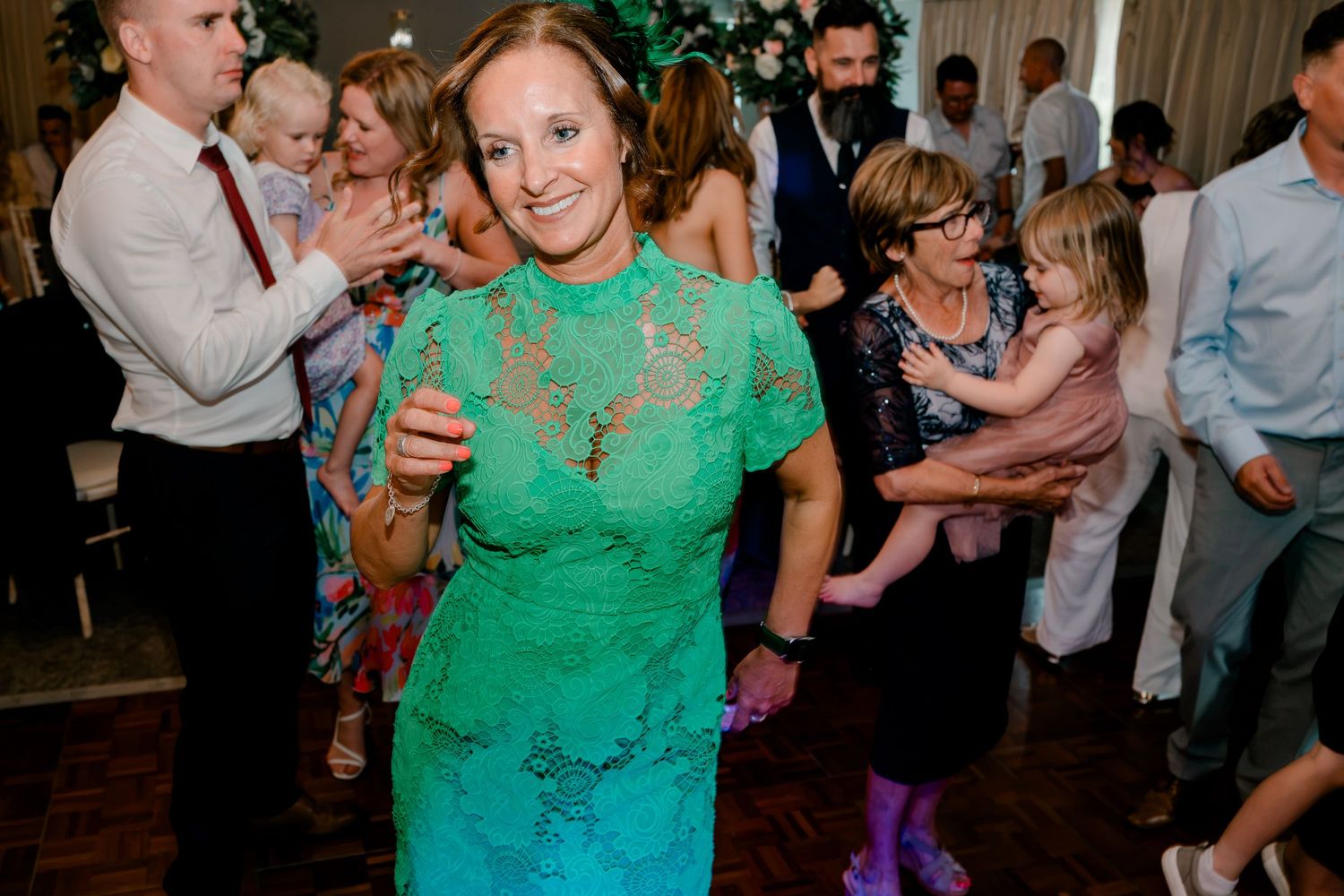 Smiling woman in a turquoise lace dress dances at a lively wedding reception as guests and children celebrate around her.