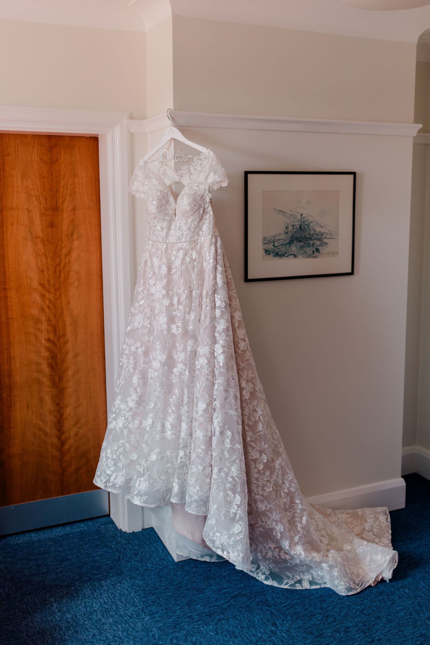 A lace wedding dress hangs elegantly against a wall with wooden doors and blue carpeting.