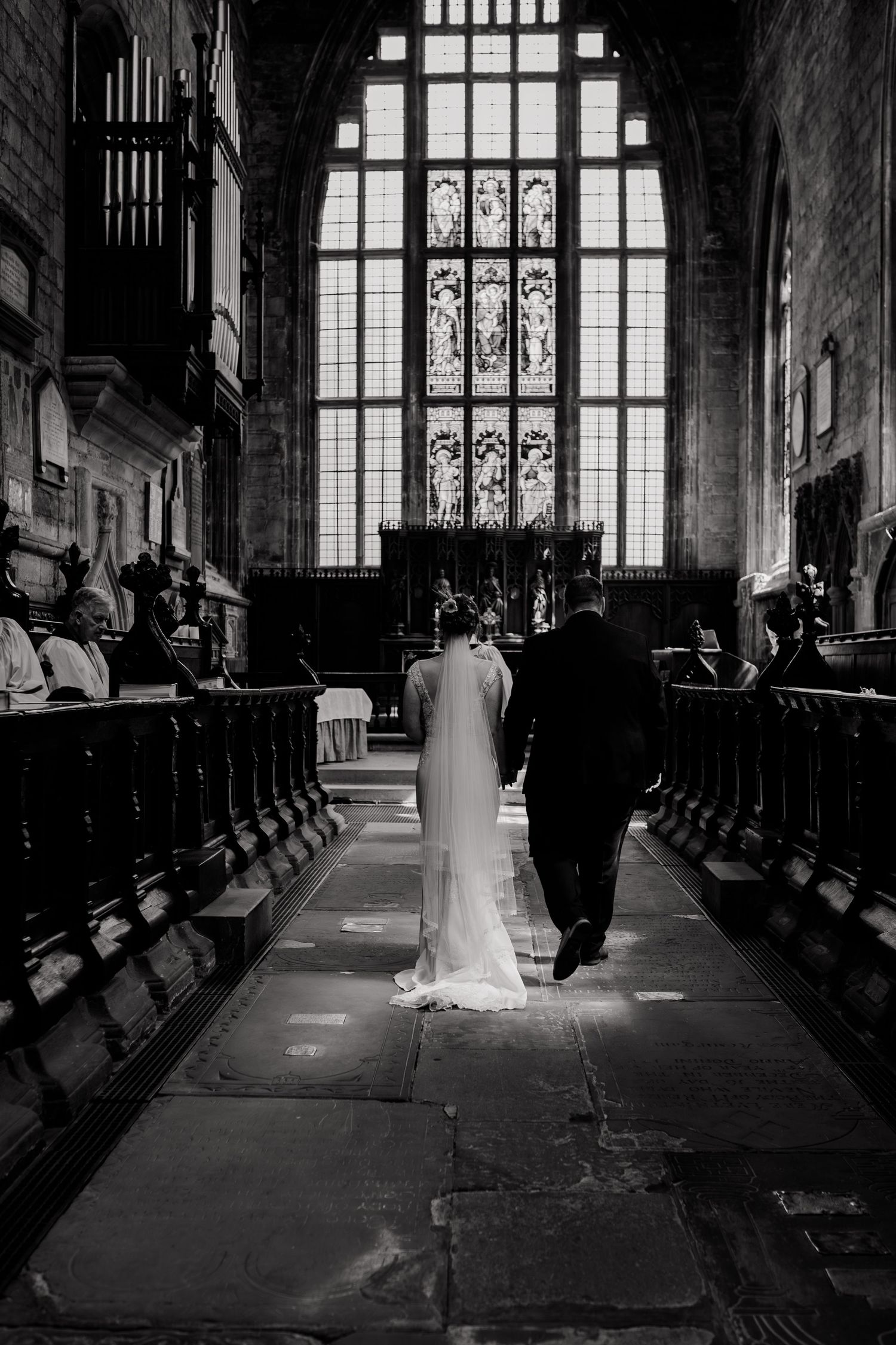 A couple walks down the aisle of a gothic church with dramatic stained glass windows and wooden pews.