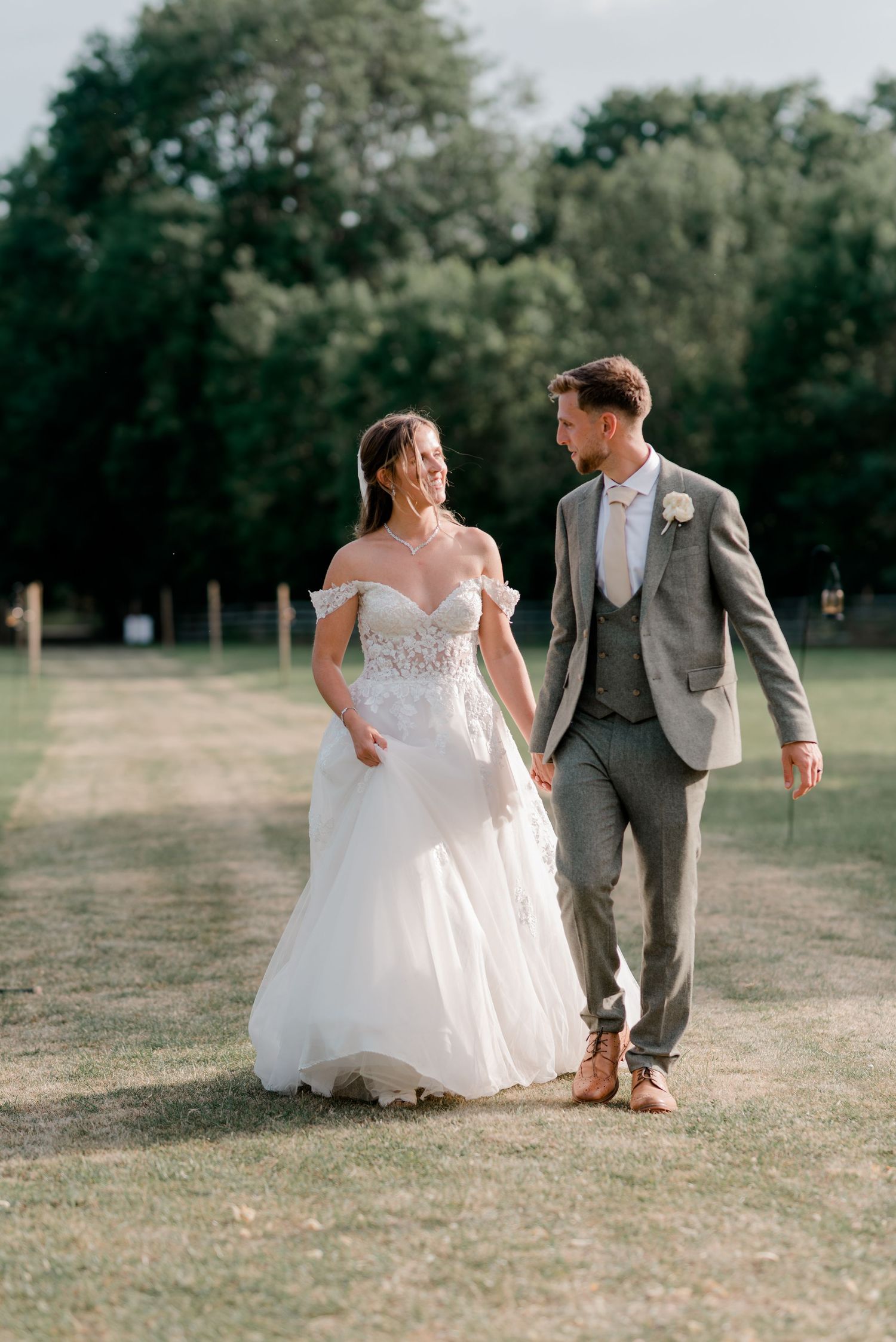 A bride in an elegant white gown and groom in gray suit walk hand in hand across a sunny outdoor venue.