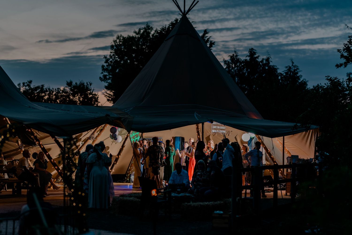 A tipi-style tent illuminated at dusk during an outdoor wedding reception with guests gathered inside.