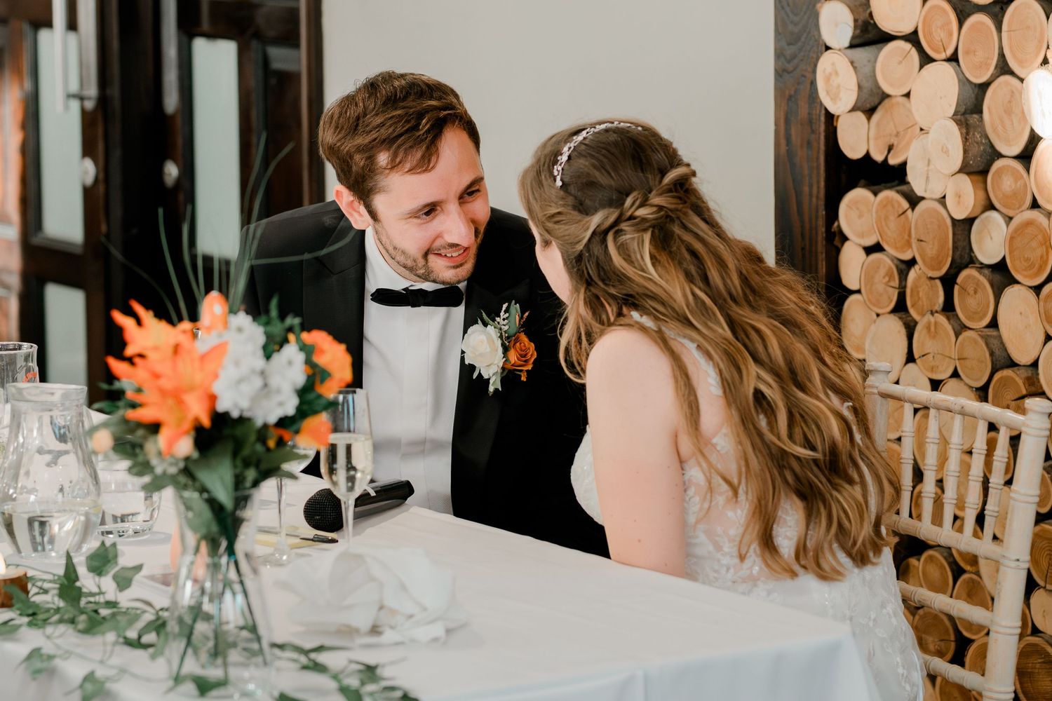Wedding reception guests enjoy dinner at elegant tables decorated with floral centerpieces and stacked wood wall accents.