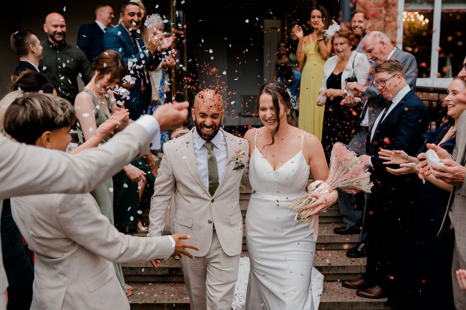Wedding guests throw confetti over a happy couple exiting their ceremony in light colored attire.