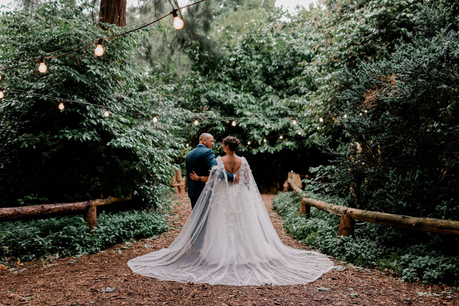 Bride in stunning cape gown walks with groom through a lush forest.