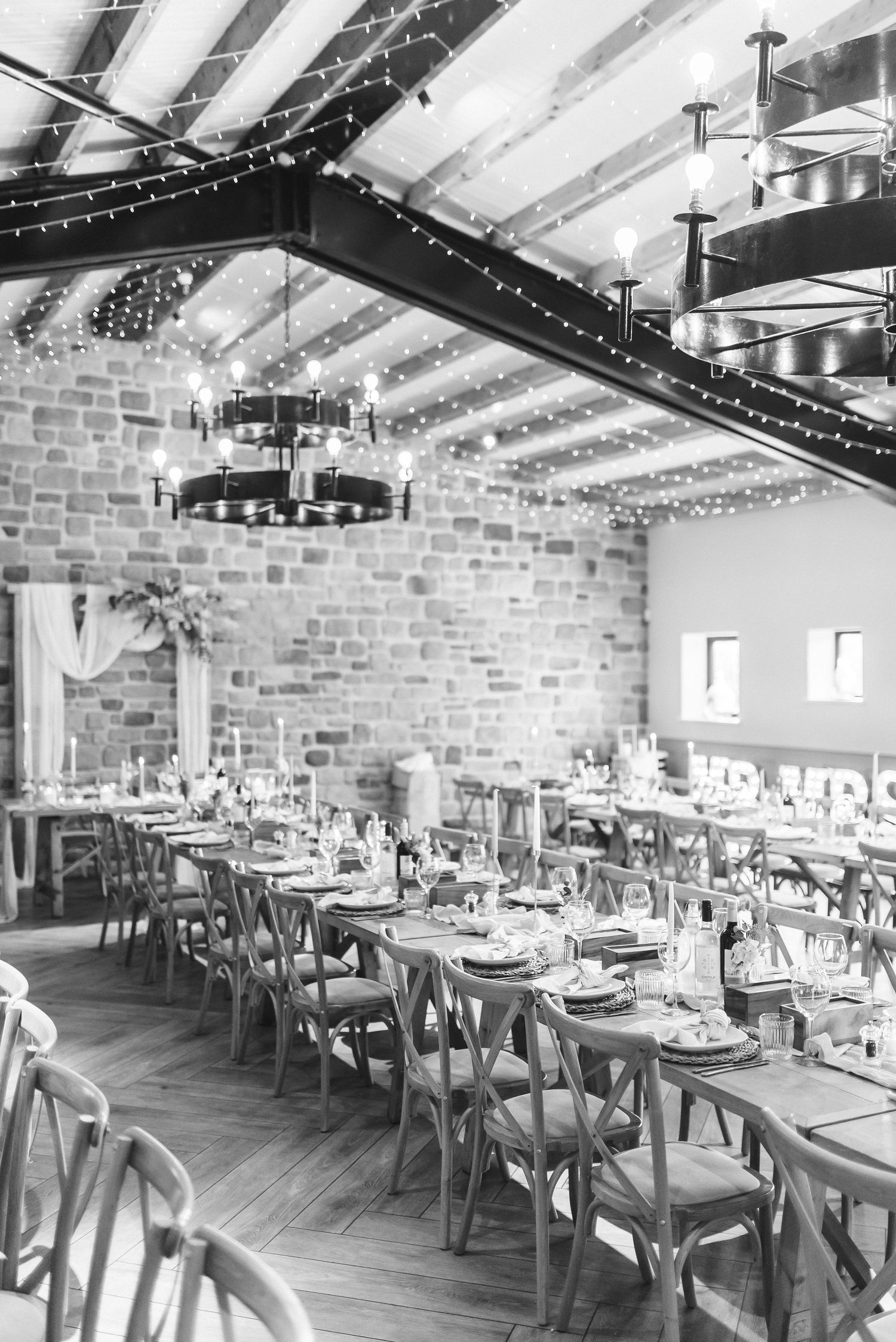 Black and white barn wedding venue interior with exposed brick, fairy lights, chandeliers and long reception tables.