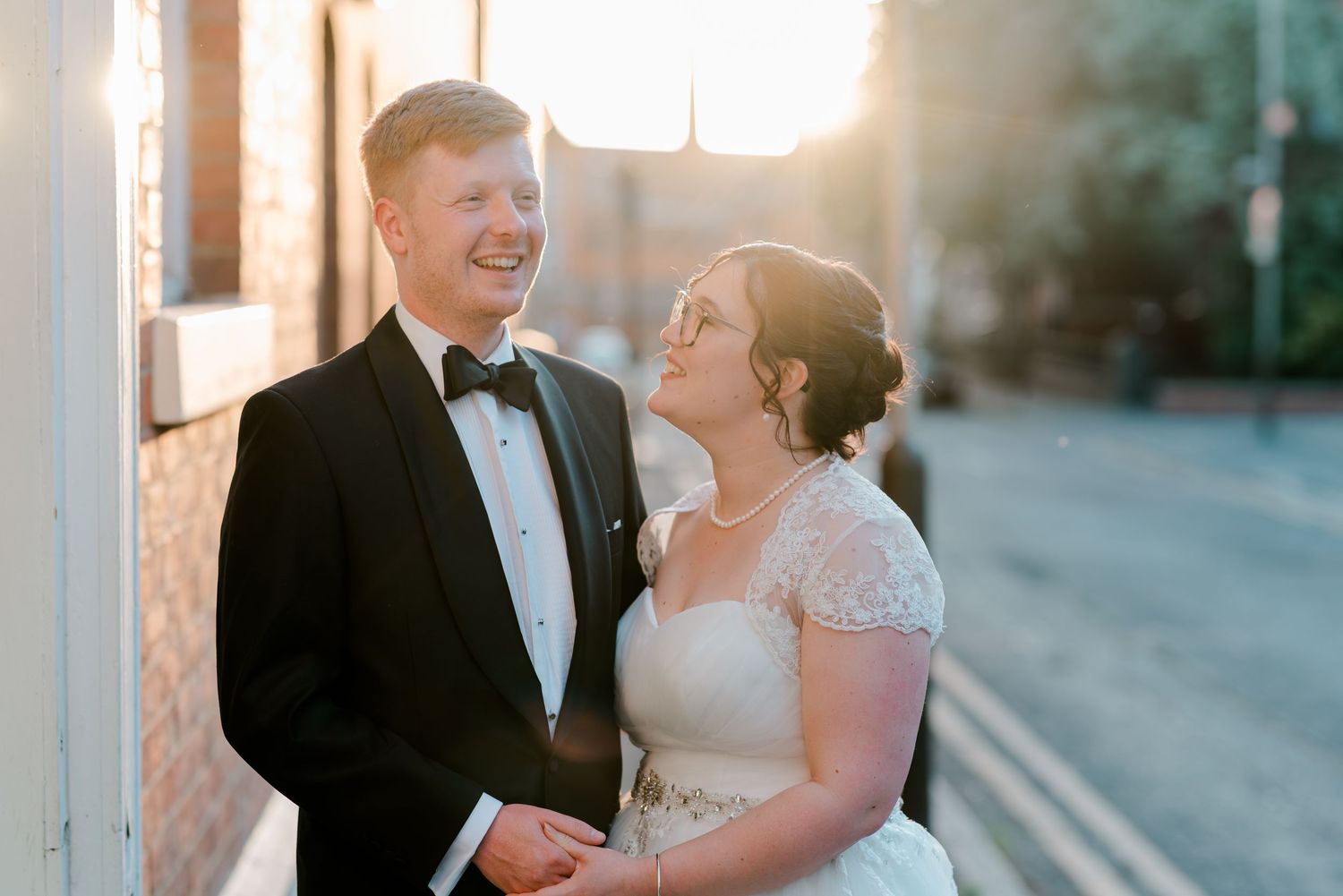 A joyful bride and groom share a laugh outdoors at golden hour, dressed in a wedding gown and black tuxedo.
