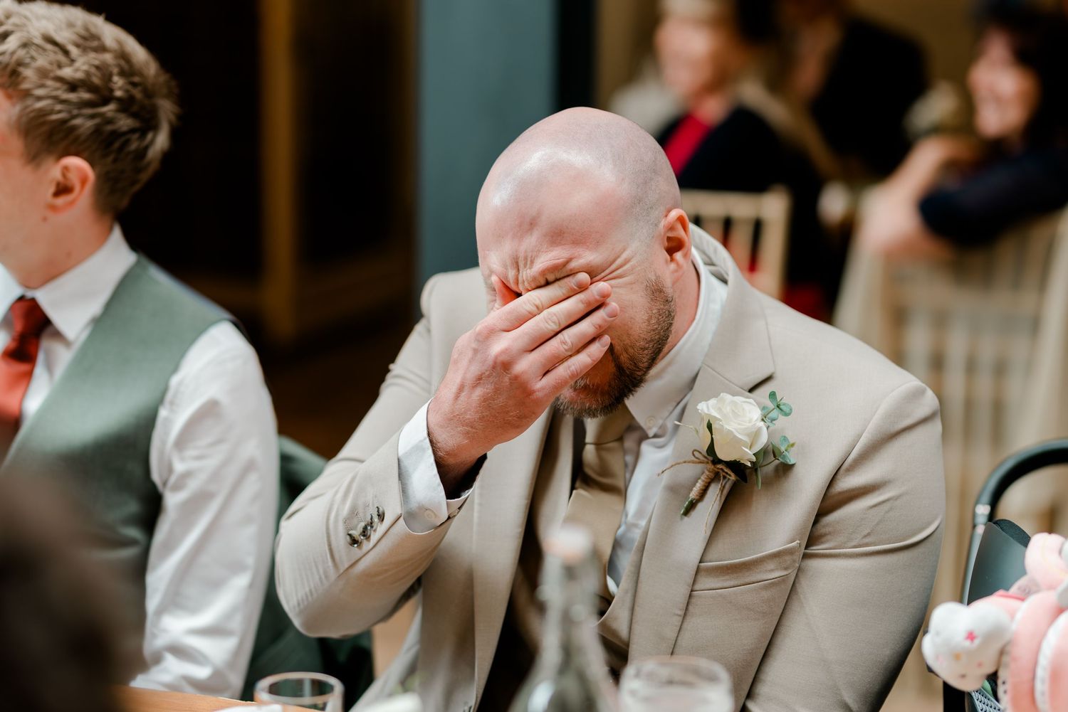 Emotional groom in beige suit covering face with hand during wedding reception speech, wearing floral boutonniere.