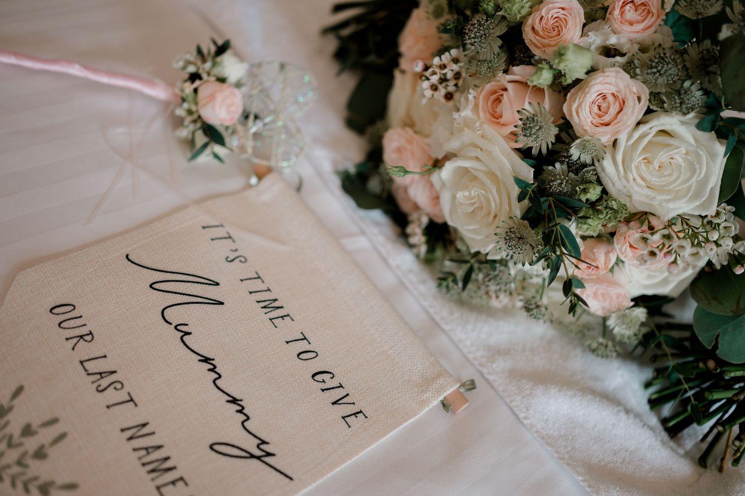 Wedding sign reading It's time to give Mummy our last name beside a pink and cream rose bouquet with greenery and a floral wrist corsage.
