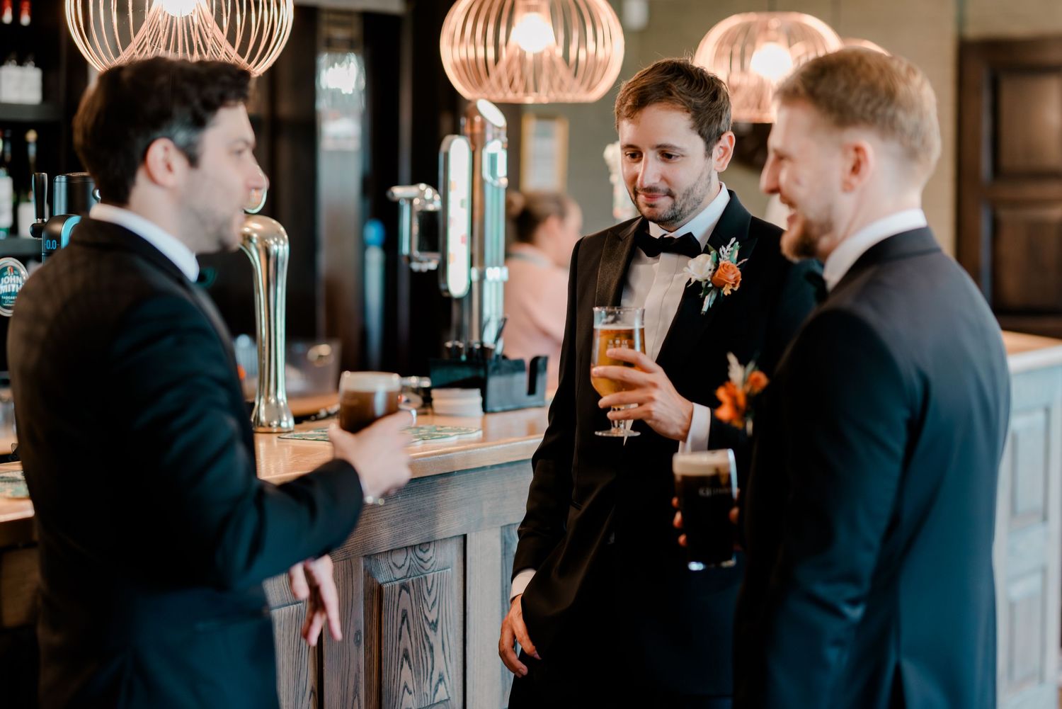 Three people in formal black suits share drinks and conversation at an elegant bar with copper pendant lighting.