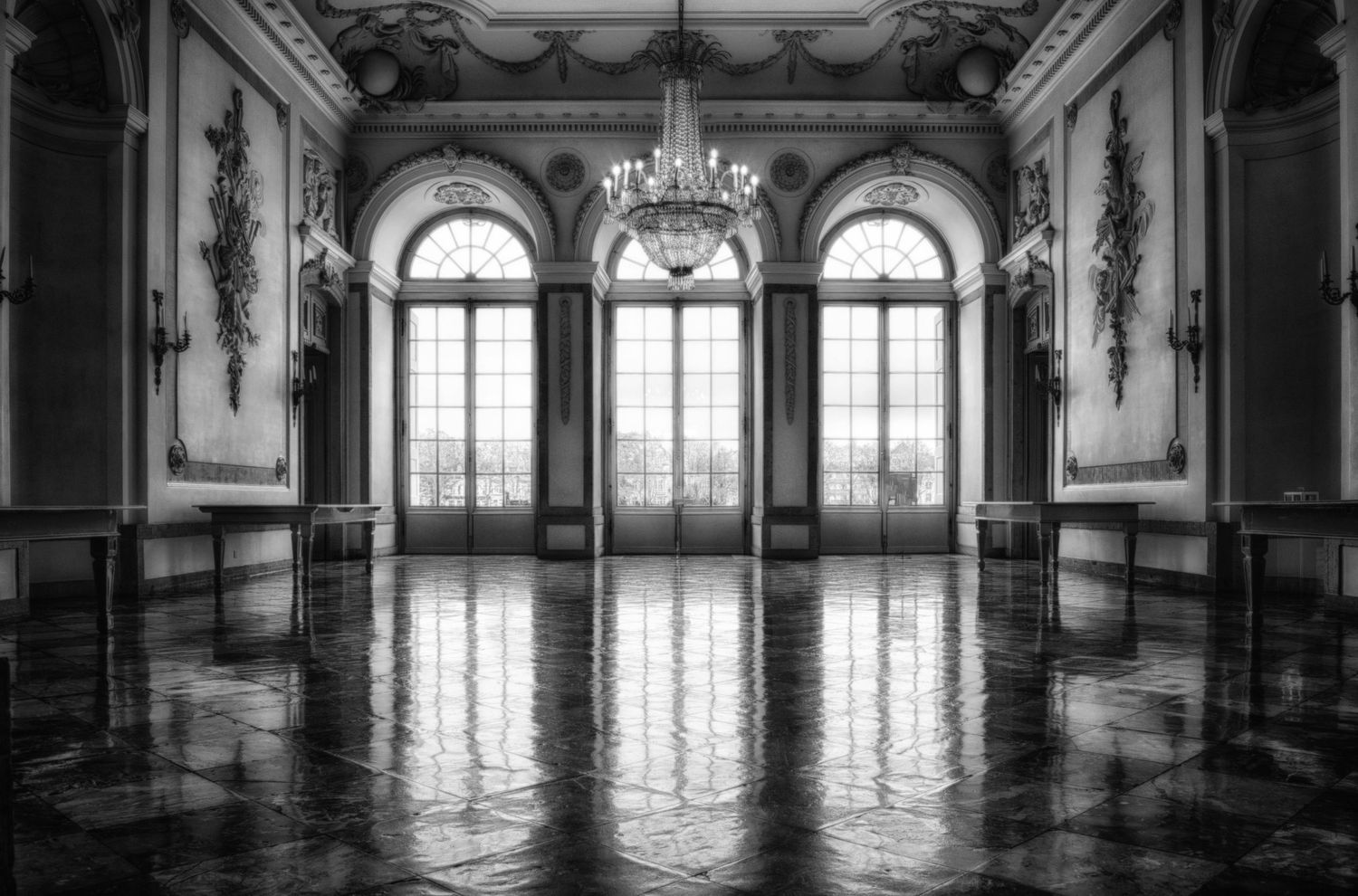 Grand ballroom with ornate chandelier and arched windows reflecting on polished marble floors.
