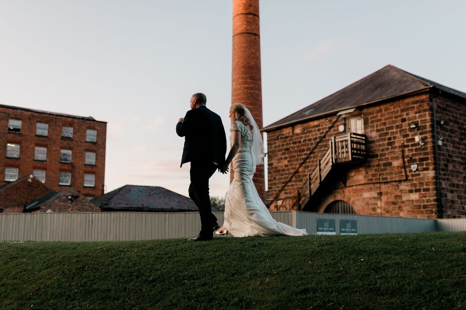 Silhouetted couple against industrial brick buildings and chimney at sunset.