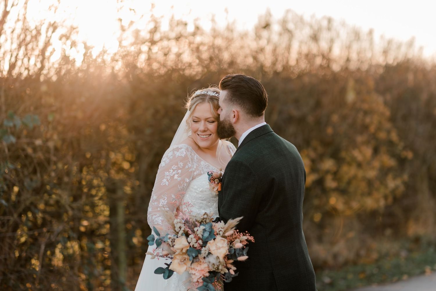 Smiling bride in lace veil holds colorful bouquet as groom gazes at her in warm autumn sunlight.