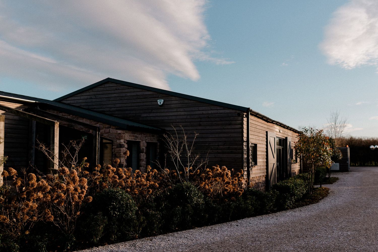 Rustic dark wood barn wedding venue exterior at dusk with gravel path and autumn wildflower garden borders.