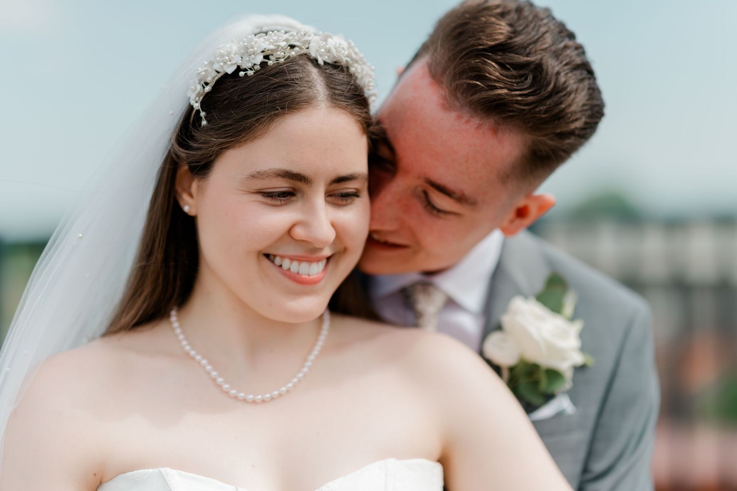 Happy bride in white gown and pearl necklace with groom in gray suit outdoors.