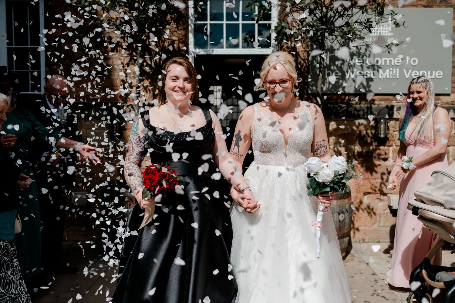 Two brides walk hand-in-hand through a shower of white confetti on their wedding day.