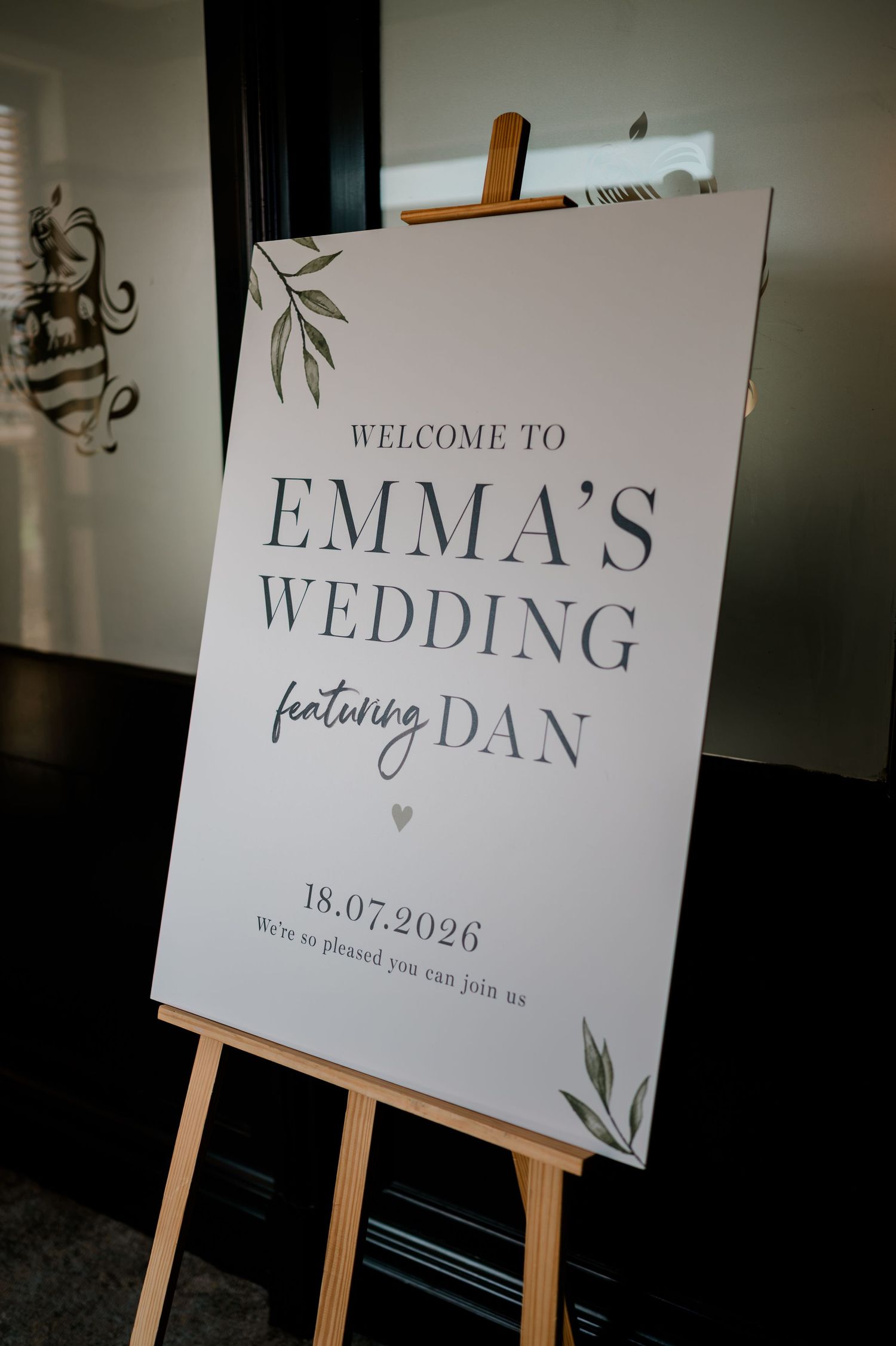 Wedding welcome sign on a wooden easel reading Emma's Wedding featuring Dan, dated 18.07.2026, with olive leaf accents.