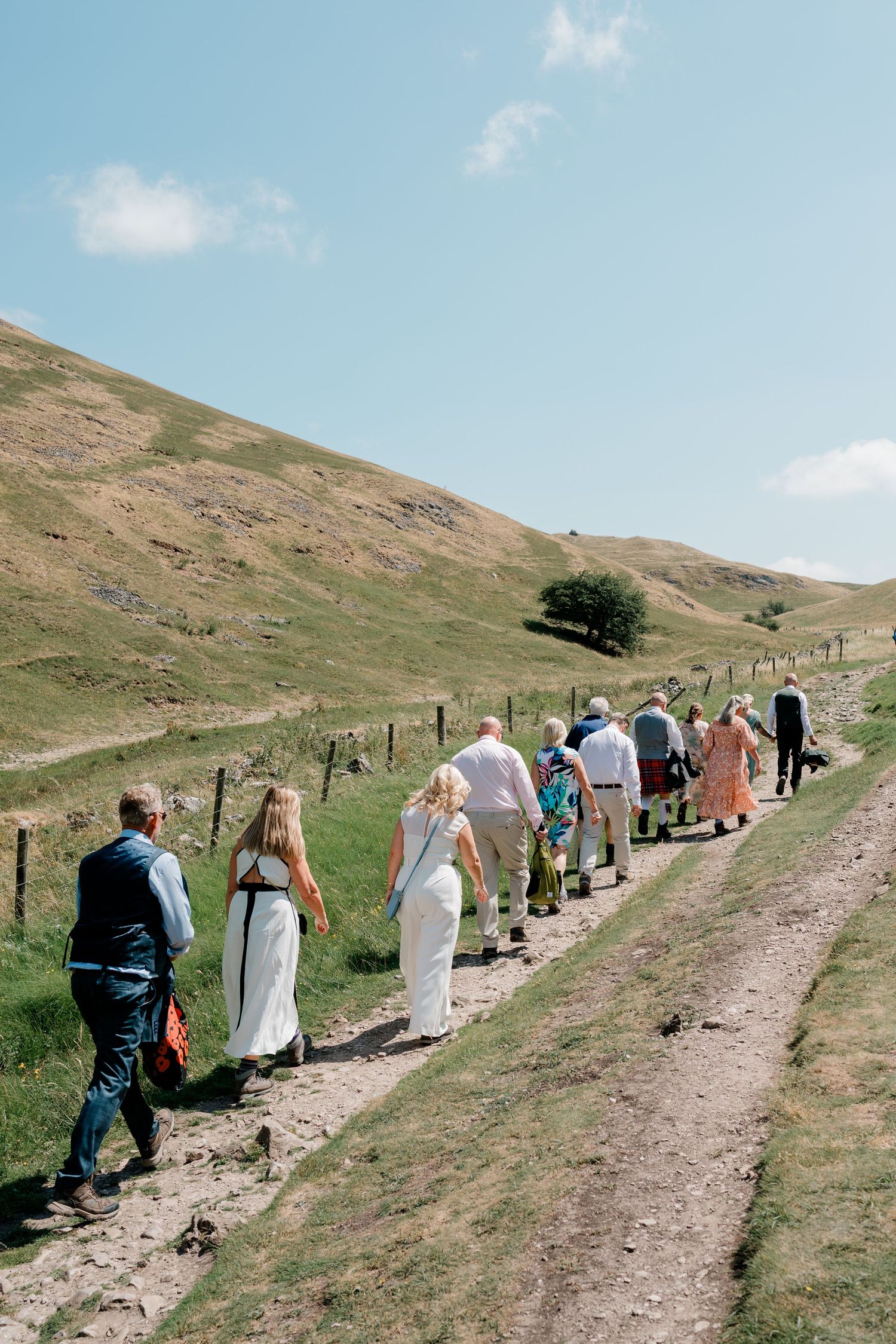 Wedding guests walk in a line along a scenic rural hillside path under a bright blue sky.