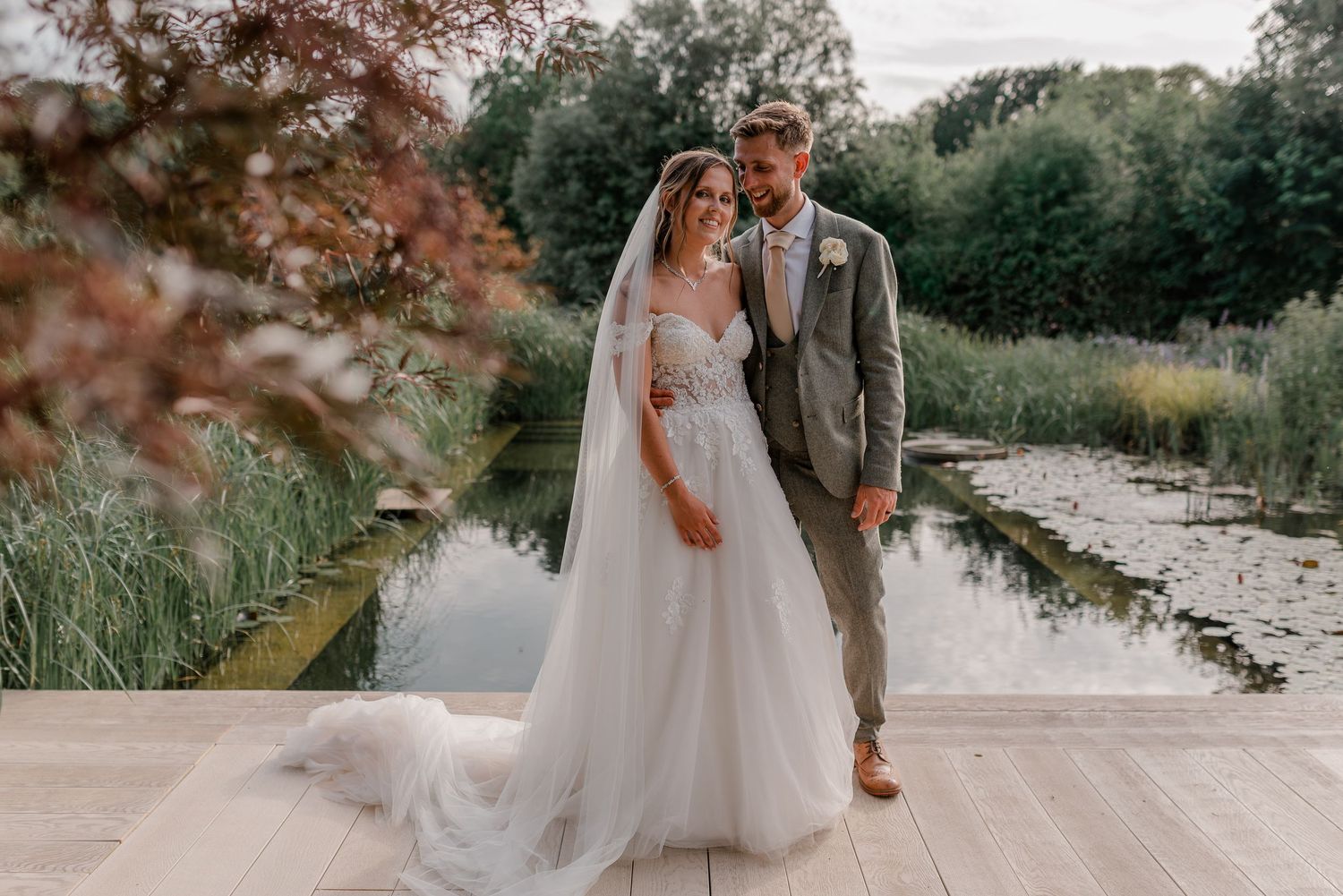 Bride in flowing tulle gown and groom in grey suit pose elegantly beside a serene garden pond.
