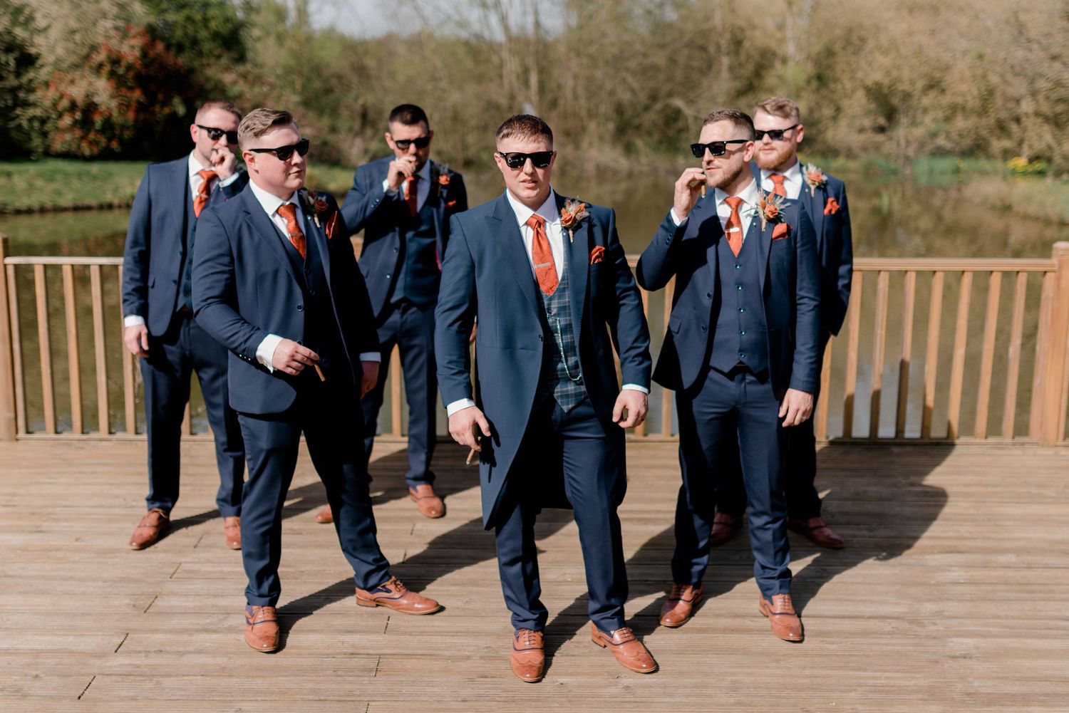 Wedding party of groomsmen in navy suits jump in unison on wooden deck showing colorful socks at outdoor venue.