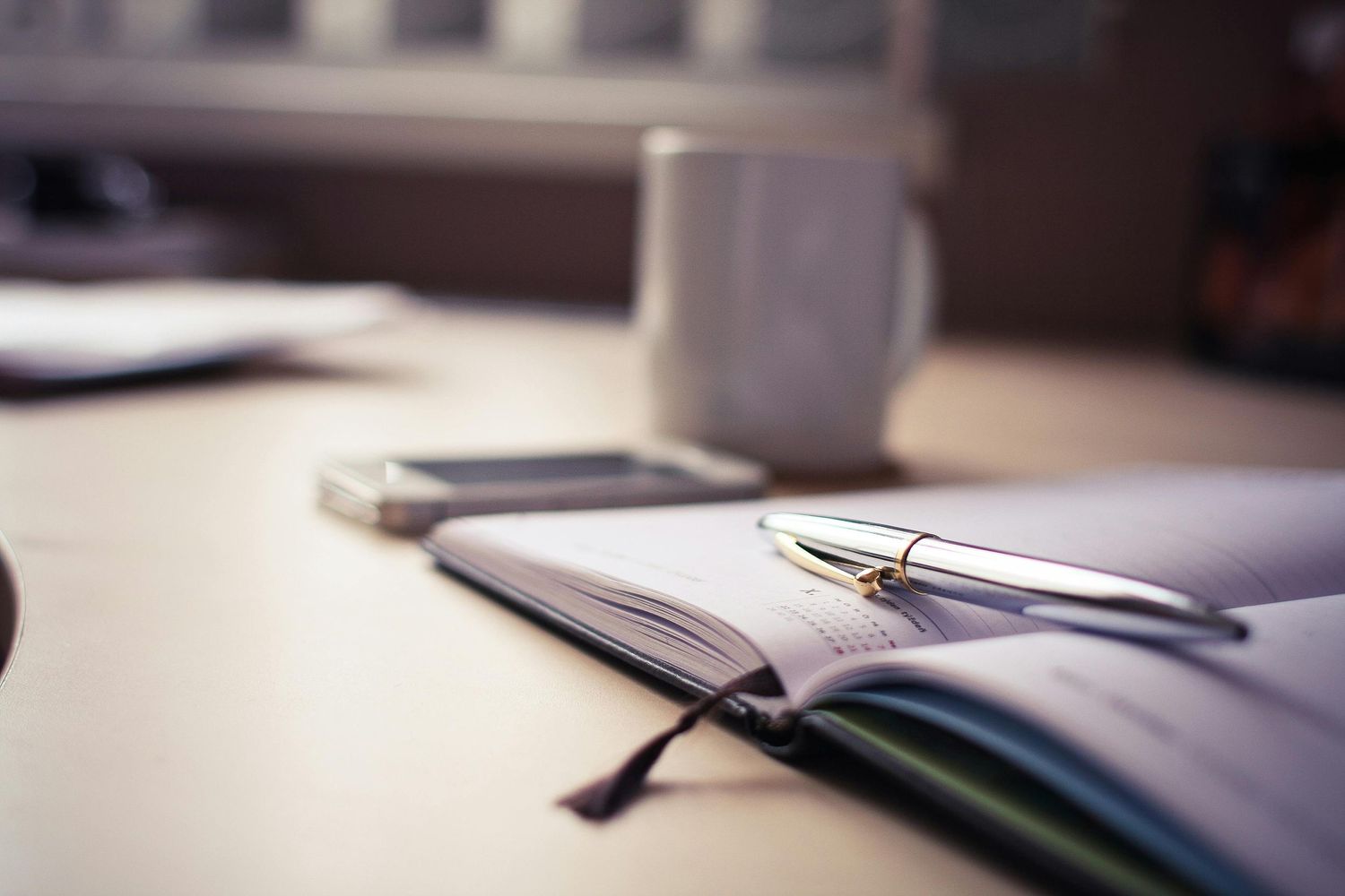 Office desk scene with notebook, pen, coffee mug and natural lighting.