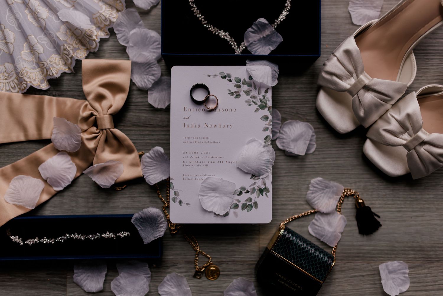 Wedding details flatlay featuring beige shoes, jewelry, invitation cards, and decorative elements on a gray wooden surface.