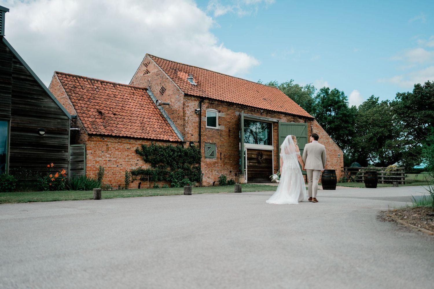 Bride and groom walking hand in hand toward a rustic red brick barn wedding venue on a sunny day.