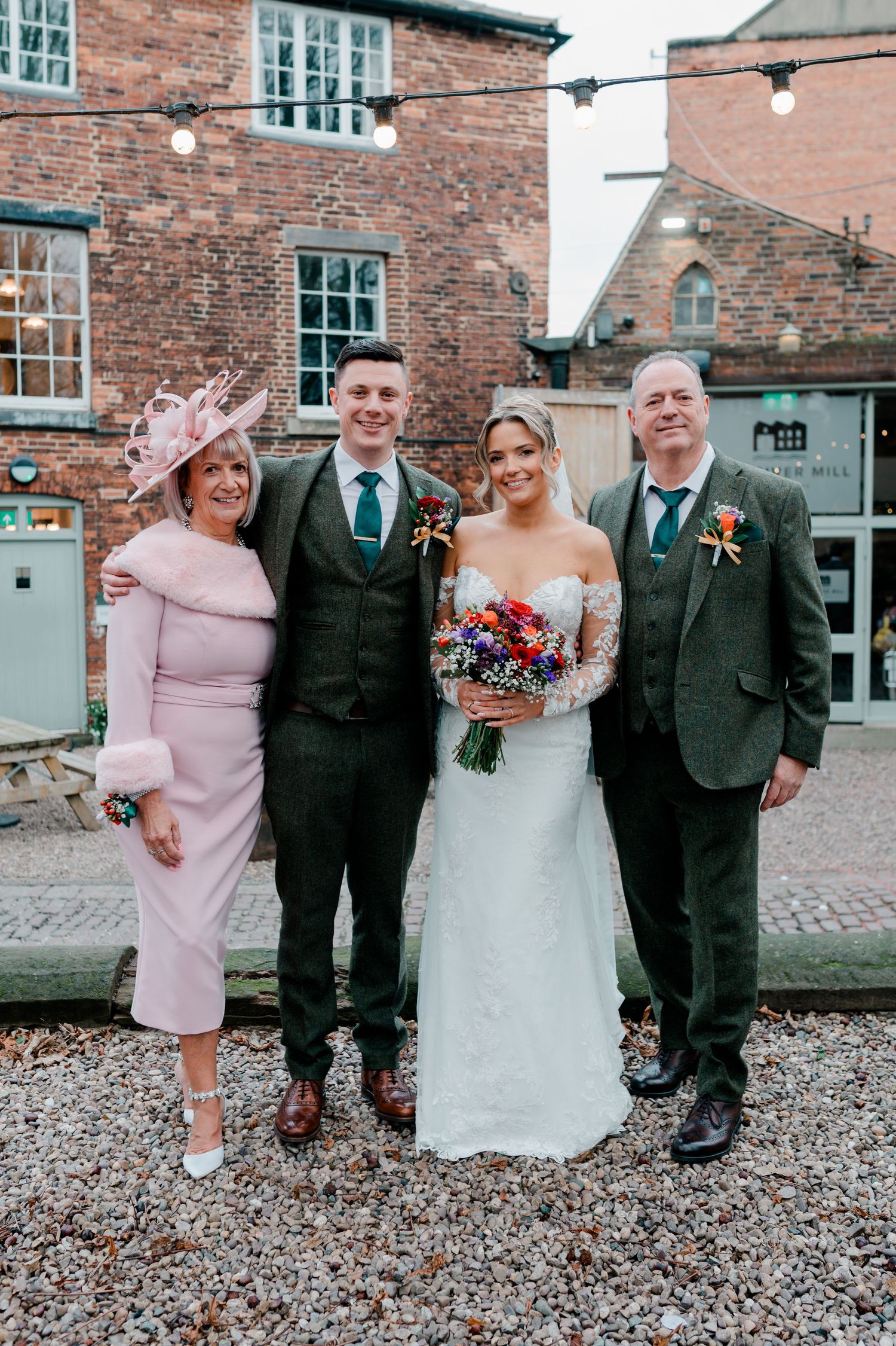 Bride and groom pose with family outside a rustic brick wedding venue with festive lights.