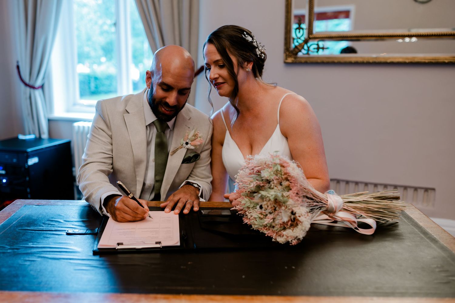 Couple signs marriage documents at a wooden table during intimate wedding ceremony.