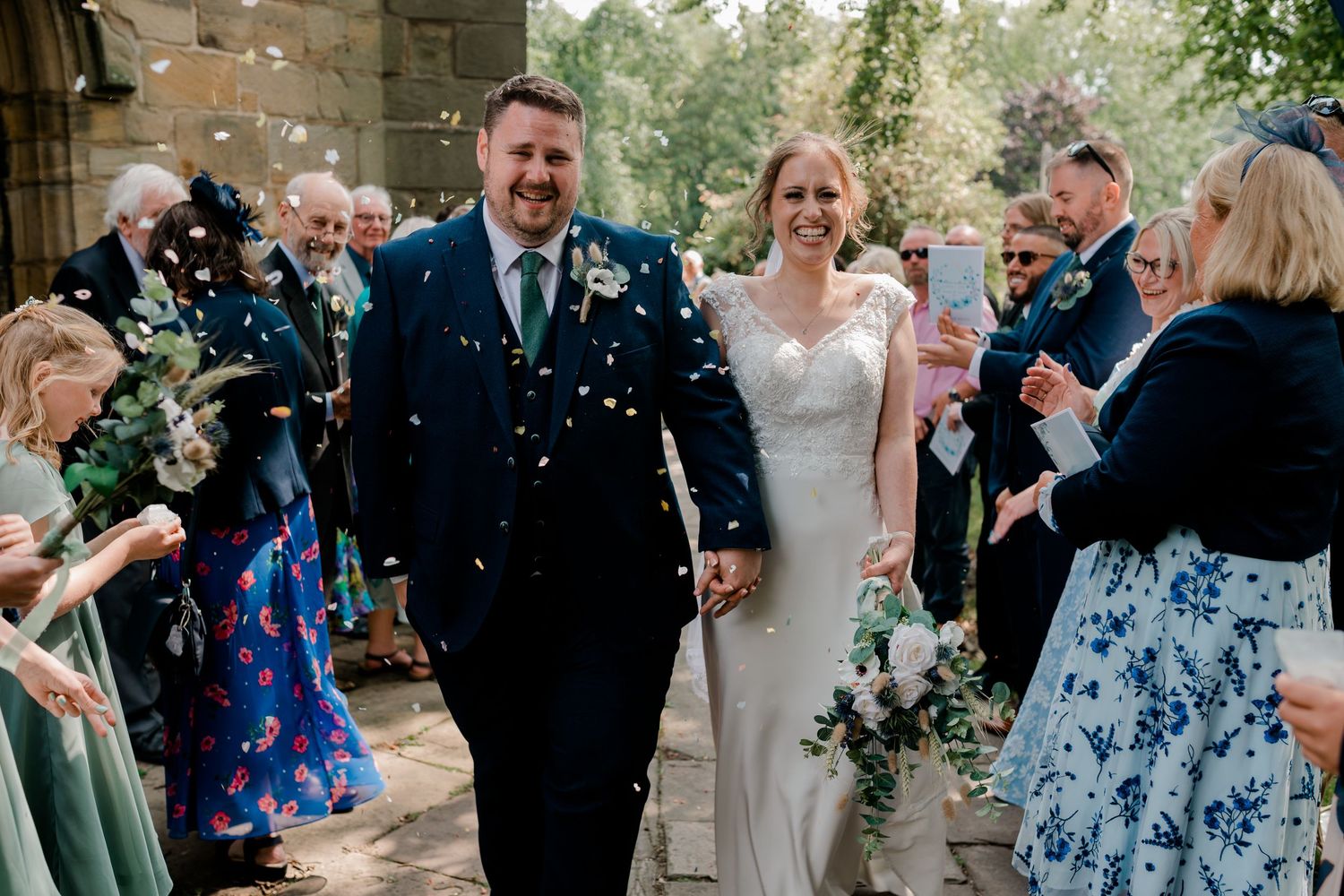 Wedding guests toss confetti in celebration outside a stone church building on a sunny day.