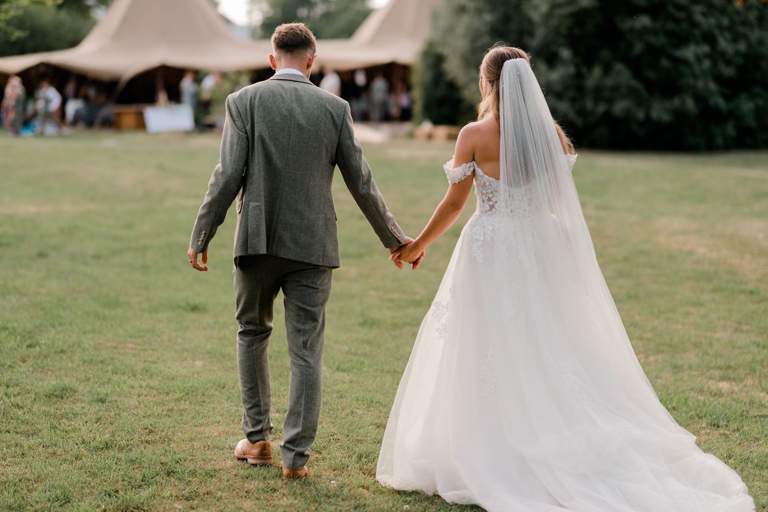 A couple in wedding attire walks hand in hand across a green lawn on their special day.