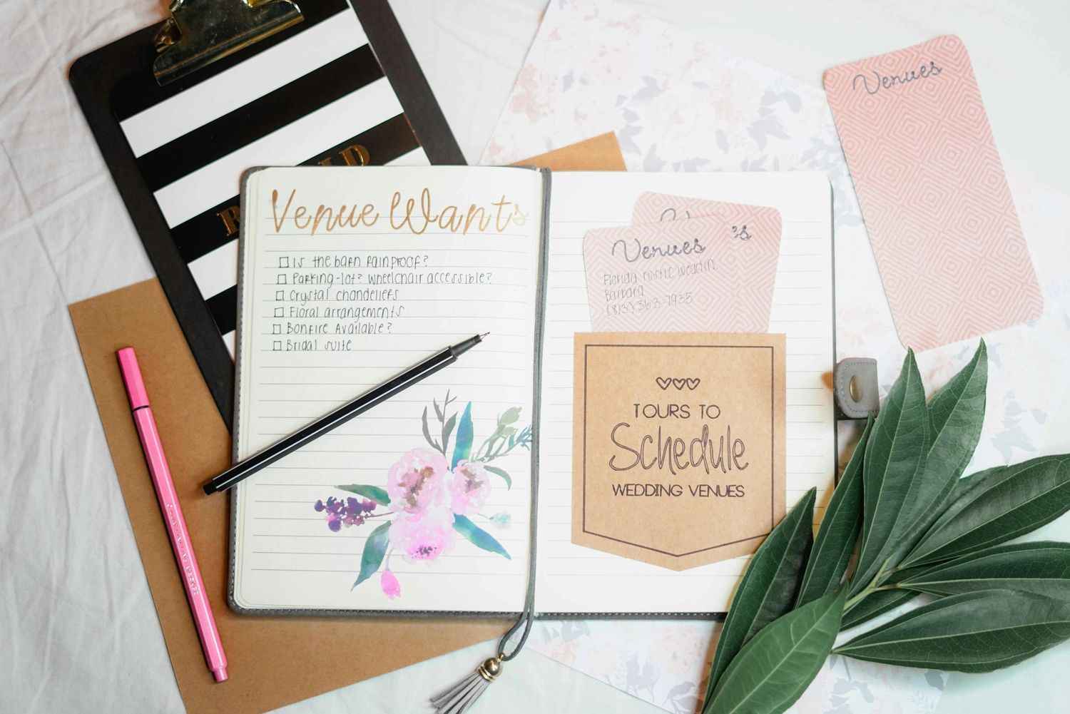 Wedding planning flatlay with floral stationery, notebook and green foliage on white surface.