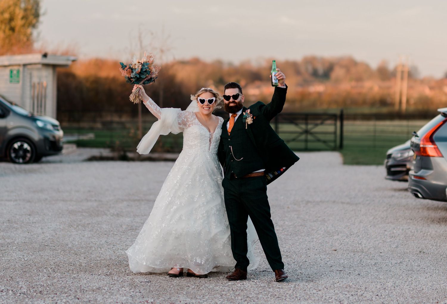 Joyful bride and groom celebrating outdoors in wedding attire, raising drinks and bouquet on a gravel venue driveway.