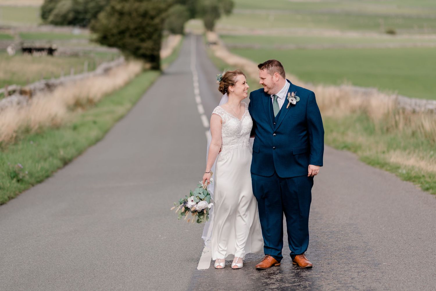 Bride and groom sharing a smile while walking on a quiet rural road on their wedding day.