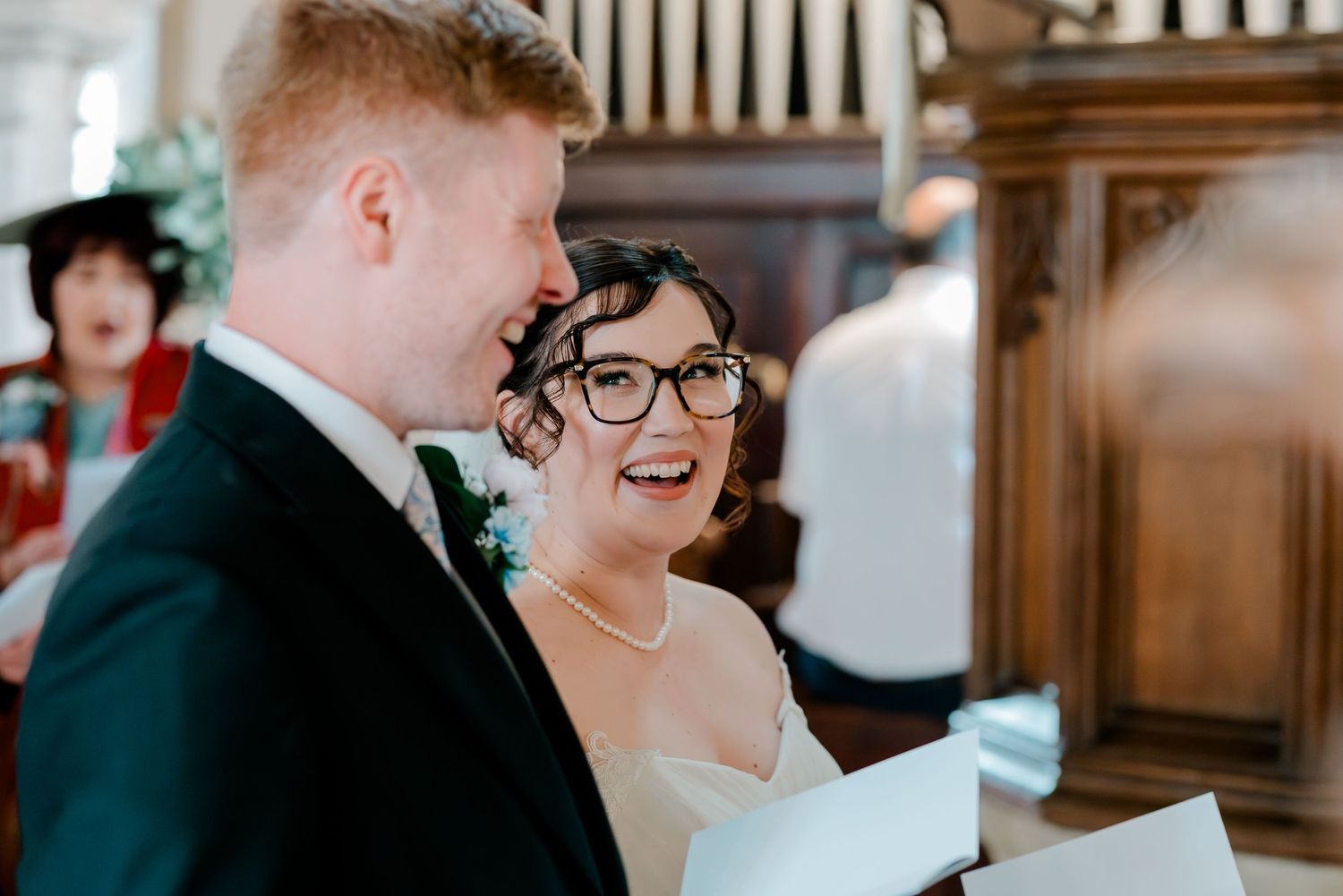 Bride and groom laughing together during wedding ceremony in a church, bride wearing glasses and pearl necklace.