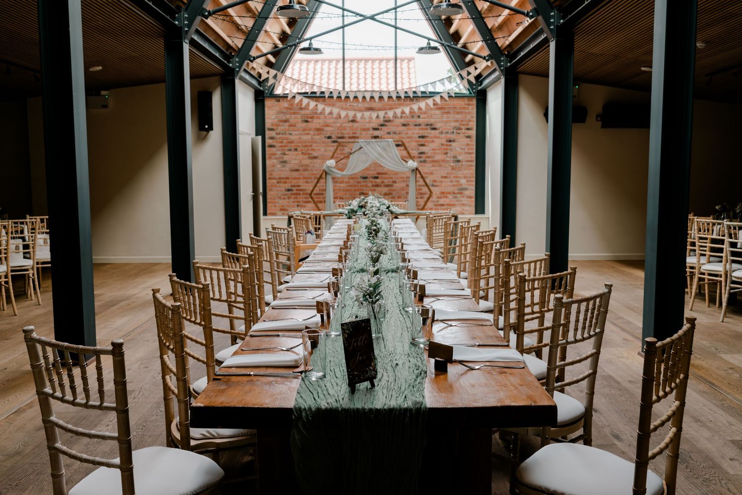 Elegant wedding reception hall with long wooden tables, gold Chiavari chairs, and lush greenery runners under a industrial skylight.